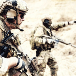 Two United States military personnel in full combat gear, one in the foreground and one in the background, in a desert-like setting. The personnel are wearing helmets with night vision goggles attached, body armor, and carrying rifles. The personnel in the foreground is in a crouching position and the personnel in the background is standing.