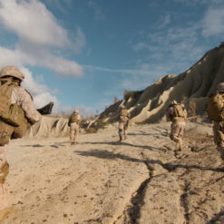 A group of US Marines in full gear walking on a dirt road in Afghanistan with a rocky cliff in the background. The Marines are carrying rifles and backpacks. The image is taken from a low angle, looking up at the Marines, and the sky is blue with a few clouds.
