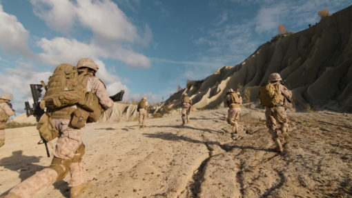 A group of US Marines in full gear walking on a dirt road in Afghanistan with a rocky cliff in the background. The Marines are carrying rifles and backpacks. The image is taken from a low angle, looking up at the Marines, and the sky is blue with a few clouds.