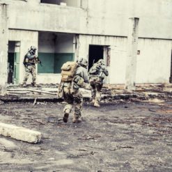 Three US military personnel in full combat gear moving through a dilapidated building in a combat zone. The personnel are wearing helmets, body armor, and carrying rifles. The building is in a state of disrepair with rubble and debris scattered about. The personnel are moving in a tactical formation with one in the lead and two following behind. The image is in a sepia tone, depicting a realistic scene of US military personnel in action.