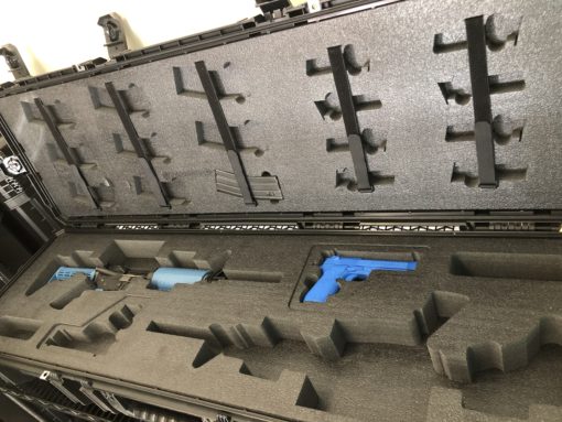 A black Pelican case with gray foam inserts for a handgun and accessories. The case is open and the handgun is blue. The foam inserts are cut to fit the handgun and accessories. The case has two latches on the front and a handle on the top. The background is a gray metal shelf.