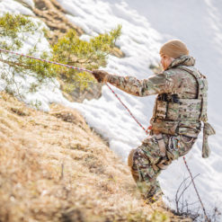 A US Army soldier in camouflage is holding onto a rope and walking down a snowy hillside. The soldier is wearing a backpack and carrying a rifle. The background consists of a hillside with trees and snow.
