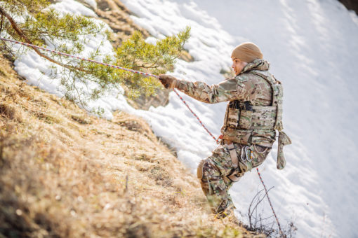 A US Army soldier in camouflage is holding onto a rope and walking down a snowy hillside. The soldier is wearing a backpack and carrying a rifle. The background consists of a hillside with trees and snow.