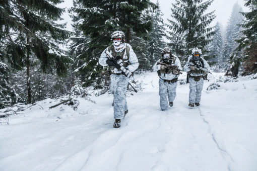 Three United States military personnel in winter camouflage walking through a snowy forest during a training exercise. The personnel are wearing white and gray camouflage clothing and carrying weapons. The background consists of a snowy forest with tall trees. The personnel are walking in a line, with the first person in the foreground and the other two following behind.