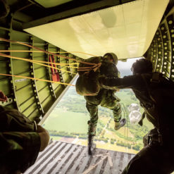 Image of military personnel parachuting from the open door of an aircraft. The aircraft is green and the personnel are wearing green uniforms and helmets. The personnel are attached to the aircraft with cords and are in the process of jumping out of the open door. The background consists of a green landscape and blue sky. The image is taken from inside the aircraft looking out towards the open door.