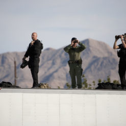 Two military personnel on a rooftop with binoculars, one holding a rifle, with mountains in the background. The personnel are wearing black and green uniforms. The rooftop appears to be white and has a security camera on the left side of the image.