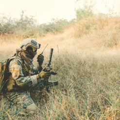 A soldier in camouflage clothing and gear kneeling in a field with tall grass and trees in the background. The soldier is holding a rifle and appears to be communicating on a radio. The soldier is wearing a helmet with a camouflage cover and a backpack.