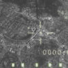 A black and white aerial view of a city with a military targeting overlay. The overlay includes a crosshair, text, and green lights at the bottom. The text on the overlay reads "OVRD", "AAG", "LOCK", "TGT", "ARM", "7429", "WHOT", and "0000-01". The green lights at the bottom appear to be indicators of some kind.