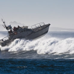 A US Coast Guard boat is seen in rough seas with waves crashing around it. The boat is white with a red stripe and has a crew on board. The boat is seen from the side and is tilted at an angle due to the waves. The waves are crashing around the boat and creating white foam. The background consists of the ocean and the horizon.