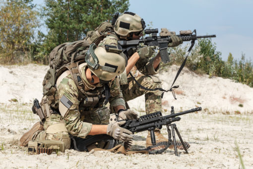 The image shows two US military personnel in full gear, holding rifles, and crouching on sandy ground. They are wearing helmets with goggles and camouflage clothing. The rifles appear to be M4 carbines with mounted scopes. The background consists of trees and shrubs, indicating a training exercise.