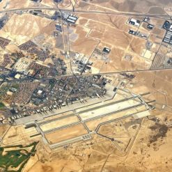 This image provides an aerial perspective of a remote military base situated in the desert. The base, enclosed by a fence, features an array of buildings and structures. The vast expanse of the desert forms the backdrop, emphasizing the base’s isolation. The base is well-developed with a network of roads and buildings, and it appears well-maintained with several vehicles and pieces of equipment visible.