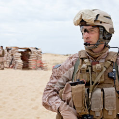 The image depicts a United States Marine in full gear standing in front of a desert training exercise. The Marine is wearing a desert camouflage uniform, a helmet, and a tactical vest with various pouches and equipment. In the background, sandbags and other Marines can be seen under clear skies.