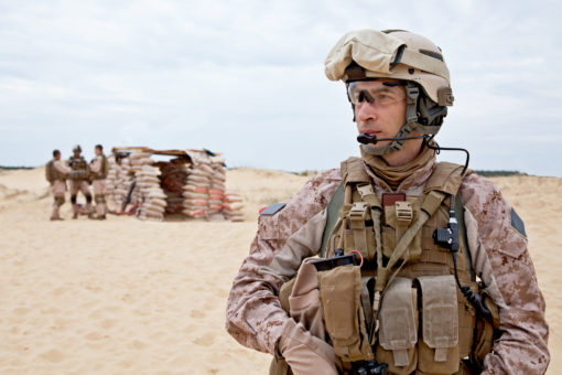 The image depicts a United States Marine in full gear standing in front of a desert training exercise. The Marine is wearing a desert camouflage uniform, a helmet, and a tactical vest with various pouches and equipment. In the background, sandbags and other Marines can be seen under clear skies.