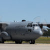 The image portrays a U.S. Air Force C-130 Hercules, a four-engine turboprop military transport aircraft, stationed on a runway. The aircraft, painted in gray, displays ‘U.S. AIR FORCE’ in white on its side and bears the tail number 93-204. Its propellers are captured in motion, suggesting imminent takeoff. The backdrop features a clear blue sky and lush green trees, contrasting with the aircraft’s utilitarian design.