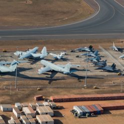 The image captures an aerial view of various types and sizes of military aircrafts, parked in a line on a curved runway. The aircrafts and runway are set against a backdrop of buildings and vehicles, all rendered in shades of brown and gray. The high-altitude perspective suggests a vast and organized military installation.