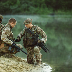 The image depicts three soldiers in camouflage gear kneeling on a riverbank, all with rifles at the ready. The soldiers are set against a backdrop of a serene river and lush trees.