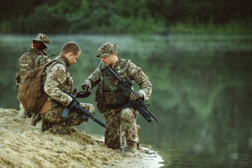 The image depicts three soldiers in camouflage gear kneeling on a riverbank, all with rifles at the ready. The soldiers are set against a backdrop of a serene river and lush trees.