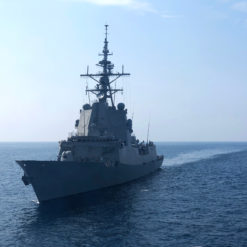 The image showcases a United States Navy Arleigh Burke-class destroyer sailing on calm sea under a clear sky. The ship, painted in gray, features a large radar tower and multiple gun turrets. The perspective is from the port side, with the ship moving towards the right side of the frame.
