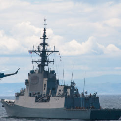 The image presents a U.S. Navy destroyer, painted in gray and equipped with a large radar tower, sailing on choppy ocean waters. A gray military helicopter is seen flying nearby, adding to the dynamic naval scene. The backdrop features the vast ocean extending towards a distant coastline under a cloudy sky.