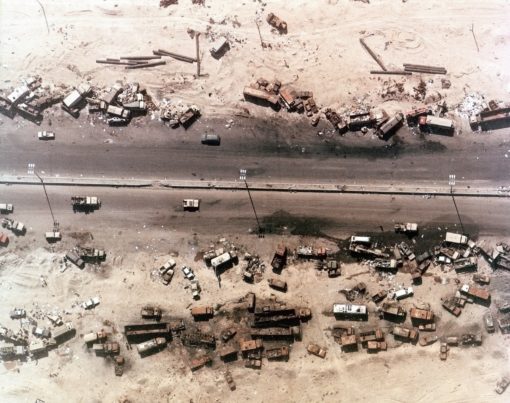The image provides an aerial perspective of a military base situated in a desert environment. The base comprises multiple rectangular buildings, possibly constructed from concrete or metal, dispersed across the sandy terrain. A variety of vehicles, including trucks and tanks, are scattered throughout the base. A road bisects the base, providing a clear path through the center. The surrounding landscape is barren, with no visible vegetation, emphasizing the isolation of the base.