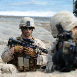 The image portrays two U.S. Marines in full gear, crouching on a rocky beach with the vast ocean as their backdrop. Dressed in desert camouflage uniforms and helmets, the Marine on the left holds a rifle with a scope, while the one on the right carries a rifle with a grenade launcher attachment.