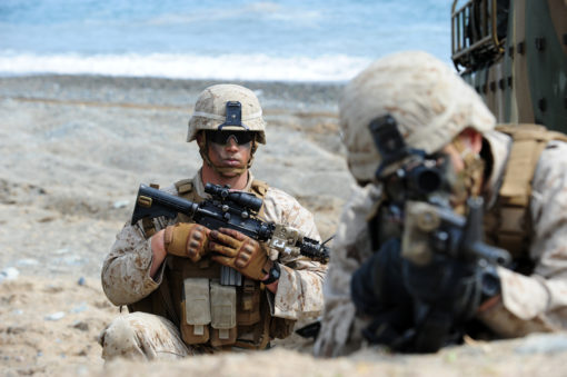 The image portrays two U.S. Marines in full gear, crouching on a rocky beach with the vast ocean as their backdrop. Dressed in desert camouflage uniforms and helmets, the Marine on the left holds a rifle with a scope, while the one on the right carries a rifle with a grenade launcher attachment.