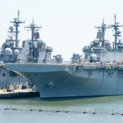 This image depicts a large, gray aircraft carrier docked in a harbor. The ship features multiple levels and towers, equipped with an array of radar and communication equipment. It is docked at a pier with several smaller boats visible in the water in front of it. The background reveals a clear blue sky and a distant city skyline.