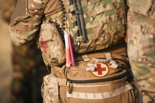 The image provides a close-up view of the torso of a military personnel, who is wearing a camouflage-patterned uniform. Attached to the uniform is an array of medical equipment, including a patch with a red cross, a tourniquet, and a pair of scissors, indicating the individual’s role in providing medical aid. The personnel is also equipped with a rifle, suggesting readiness for combat. The blurred background hints at an outdoor setting, possibly a field or training exercise. This image underscores the multifaceted roles and responsibilities within the military.