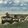 This image portrays a convoy of three tan-colored military vehicles driving on a dirt road through a grassy field. The first vehicle, marked with the number ‘13’, is the largest and features a turret on top. The second is a smaller truck with a mounted gun, and the third is a larger truck with a covered cargo area. The vehicles are kicking up a large cloud of dust behind them. In the background, a blue sky with white clouds and distant wind turbines can be seen.