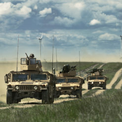 This image portrays a convoy of three tan-colored military vehicles driving on a dirt road through a grassy field. The first vehicle, marked with the number ‘13’, is the largest and features a turret on top. The second is a smaller truck with a mounted gun, and the third is a larger truck with a covered cargo area. The vehicles are kicking up a large cloud of dust behind them. In the background, a blue sky with white clouds and distant wind turbines can be seen.