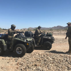This image shows three soldiers in camouflage gear riding on two all-terrain vehicles (ATVs) in a desert setting. The ATVs are black and green. The soldiers, equipped with helmets and backpacks, navigate the rocky desert landscape under a clear, blue sky. Mountains can be seen in the distance.