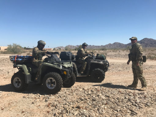 This image shows three soldiers in camouflage gear riding on two all-terrain vehicles (ATVs) in a desert setting. The ATVs are black and green. The soldiers, equipped with helmets and backpacks, navigate the rocky desert landscape under a clear, blue sky. Mountains can be seen in the distance.