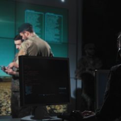 This image captures a moment during a military briefing in a high-tech room. The room is dimly lit, enhancing the large screen in the background that displays an image of a rocket launch. Two individuals are in the foreground, one seated at a desk with a computer monitor and wearing a headset, possibly indicating their role as a communications officer. The other individual is standing, holding a clipboard, and appears to be giving instructions, suggesting they may be the team leader or coordinator. A third person stands near the screen in the background, possibly observing or waiting their turn to present.