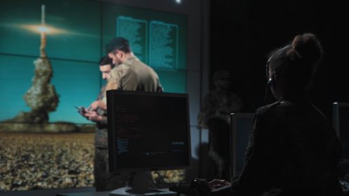 This image captures a moment during a military briefing in a high-tech room. The room is dimly lit, enhancing the large screen in the background that displays an image of a rocket launch. Two individuals are in the foreground, one seated at a desk with a computer monitor and wearing a headset, possibly indicating their role as a communications officer. The other individual is standing, holding a clipboard, and appears to be giving instructions, suggesting they may be the team leader or coordinator. A third person stands near the screen in the background, possibly observing or waiting their turn to present.