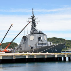 A large naval ship, painted in gray and marked with the number ‘174’, is docked at a concrete pier with a yellow railing. The ship features a prominent tower, equipped with multiple antennas and radar dishes, indicative of its advanced communication and surveillance capabilities. Adjacent to the ship, two large cranes stand on the pier. The background reveals a clear blue sky and a tree-lined shore.
