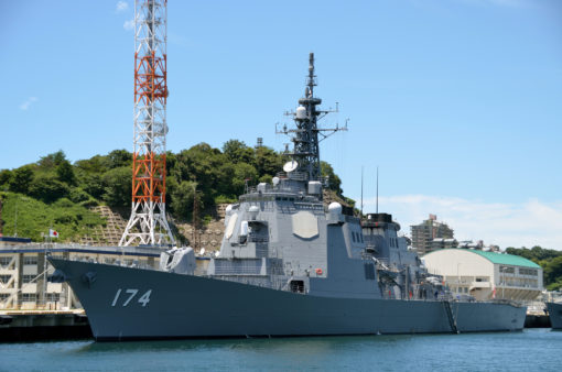 This image depicts a large, gray naval ship, numbered ‘174’, docked at a harbor. The ship features a large radar tower and multiple antennas on top. It is docked next to a red and white tower crane, with a green hill populated by trees nearby. The water around the ship is calm under a blue sky with some clouds. Some buildings are visible in the background