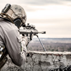 This image portrays a soldier in full gear aiming a rifle over a concrete wall. The soldier is equipped with a helmet featuring a mounted camera, and a gas mask. The rifle is fitted with a scope. The background reveals a landscape of trees and fields under a cloudy sky, creating an overall tense mood.