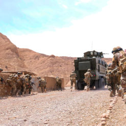 a group of soldiers patrolling a rocky, arid area with large, towering brown mountains in the background. The soldiers are wearing combat uniforms, helmets, and protective vests, carrying rifles and equipment on their backs. They are moving alongside a stone wall, with some using it for cover. Their formation suggests they are cautiously advancing in the area. On their left, an armored military vehicle, painted in a neutral color, is stationed with its antenna and turret visible. The ground is uneven, scattered with small rocks, and the sky above is blue with a hint of clouds. The overall scene conveys a sense of readiness and vigilance in a challenging environment.