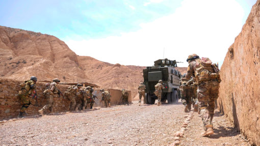a group of soldiers patrolling a rocky, arid area with large, towering brown mountains in the background. The soldiers are wearing combat uniforms, helmets, and protective vests, carrying rifles and equipment on their backs. They are moving alongside a stone wall, with some using it for cover. Their formation suggests they are cautiously advancing in the area. On their left, an armored military vehicle, painted in a neutral color, is stationed with its antenna and turret visible. The ground is uneven, scattered with small rocks, and the sky above is blue with a hint of clouds. The overall scene conveys a sense of readiness and vigilance in a challenging environment.