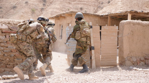 Three soldiers in full gear, wearing camouflage uniforms and helmets, are walking through a desert village. They are armed with rifles, prepared for any situation. The soldier on the left is equipped with a backpack, while the one on the right sports a vest. The background reveals a dirt road, a stone wall, and a small building with a thatched roof. The sky above is clear, and the terrain is rocky and barren, typical of a desert environment.