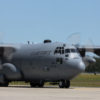 A photograph showcasing a U.S. Air Force cargo plane, specifically a C-130 Hercules, on a tarmac under a clear sky. The plane is a large, four-engine turboprop military transport aircraft, with its propellers visibly spinning. Painted in a muted gray, the words "U.S. AIR FORCE" are prominently displayed on its fuselage. The aircraft's sturdy landing gear is in contact with the concrete runway, and its characteristic high-wing design is evident. The overall atmosphere depicts a scene of readiness, with the plane either preparing for takeoff or having just landed. The backdrop is serene with an expansive blue sky.