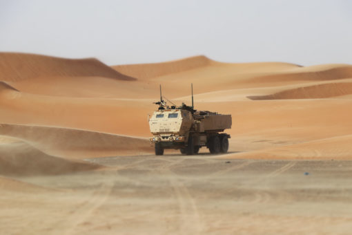 A beige military vehicle, robust and angular in design, stands prominently against a vast desert backdrop. The vehicle, equipped with multiple antennas and mounted equipment, is positioned on a tire-treaded path amidst a sea of undulating sand dunes. These dunes, characterized by their smooth, wavy patterns, transition in hue from deep tan at the bases to a lighter golden at their peaks, capturing the essence of a sunlit desert. The horizon is vast, only interrupted by the gentle curvature of the dunes and the expansive blue sky above, encapsulating the isolation and expanse of the arid environment.