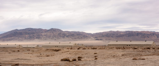A photo-realistic image of a desert landscape featuring a few long, low buildings with flat roofs and beige walls in the foreground. The foreground is mostly barren, interspersed with patches of dry grass and shrubs. An overcast sky looms above, with a few patches of blue peeking through the clouds. In the background, mountains of brown and gray rise, with patches of snow adorning their peaks.