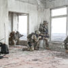 A group of six heavily-equipped soldiers in a dilapidated room with peeling paint and debris on the floor. The soldiers wear full combat gear, including helmets with mounted cameras, ballistic vests, and patches indicating U.S. affiliation. They carry modern rifles and some possess additional equipment such as radios and scopes. To the left, two soldiers are in communication: one kneeling with a radio, the other standing with a focus on the radio's transmission. In the center, three soldiers are positioned near large, weathered windows, aiming their rifles outward with disciplined stances. To the right, one soldier is crouched by a window, scanning the horizon. The windows provide a muted view of a vast, open landscape. The ambiance suggests a strategic operation in an urban environment amidst desolation.