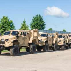 A convoy of large, boxy, tan-colored military vehicles, possibly armored, parked in a line on a paved road. The road is surrounded by grass and trees, under a blue sky with a few clouds. A red building is visible in the background.