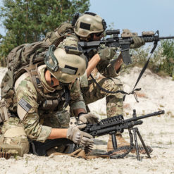 The image presents two soldiers in full combat gear, kneeling on sandy terrain and aiming their rifles. They are dressed in camouflage uniforms, helmets, and bulletproof vests. The rifles, equipped with scopes, are black. In the background, there are bushes and trees in the distance. The soldiers’ faces are blurred for privacy reasons, emphasizing the focus on their actions and equipment.