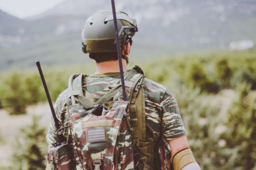 A photo-realistic image of a person in military gear, seen from behind, walking through a field. The individual is wearing a green helmet with a black strap, and a camouflage backpack equipped with a radio antenna and a rifle. The person’s camouflage uniform blends with the surrounding field scattered with trees. In the distance, mountains rise against the backdrop of the sky. The image has a faded, vintage feel to it.