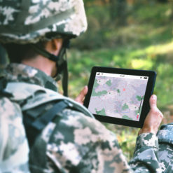 This image captures a soldier, dressed in camouflage uniform and equipped with a helmet and backpack, in a forest setting. The soldier is holding a tablet that displays a map with red markers. The perspective is from behind the soldier, looking over his shoulder, providing a clear view of the tablet screen and the surrounding trees and foliage. The image conveys a sense of strategic planning in a natural environment.