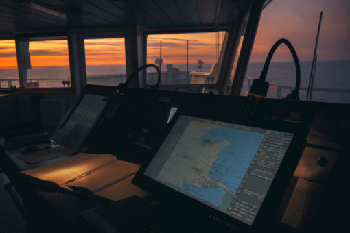 The image captures the breathtaking view from a ship’s bridge at sunset. The perspective is from the captain’s chair, showcasing modern navigational equipment such as a radar, a chart plotter displaying a map of the Florida coast, and a computer. The panoramic windows offer an unobstructed view of the vast ocean meeting an orange sky at the horizon. The bridge is devoid of personnel, adding to the tranquility of the scene.