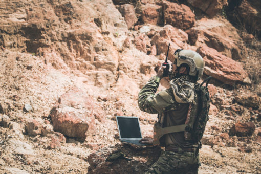 A U.S. soldier, fully equipped in camouflage gear, is kneeling on a rocky terrain. He is wearing a helmet with a headset and holding a rifle, indicating readiness for action. In front of him, an open laptop rests on a rock, suggesting communication or strategic planning. The background features a rugged cliff face, adding to the serious and tense atmosphere